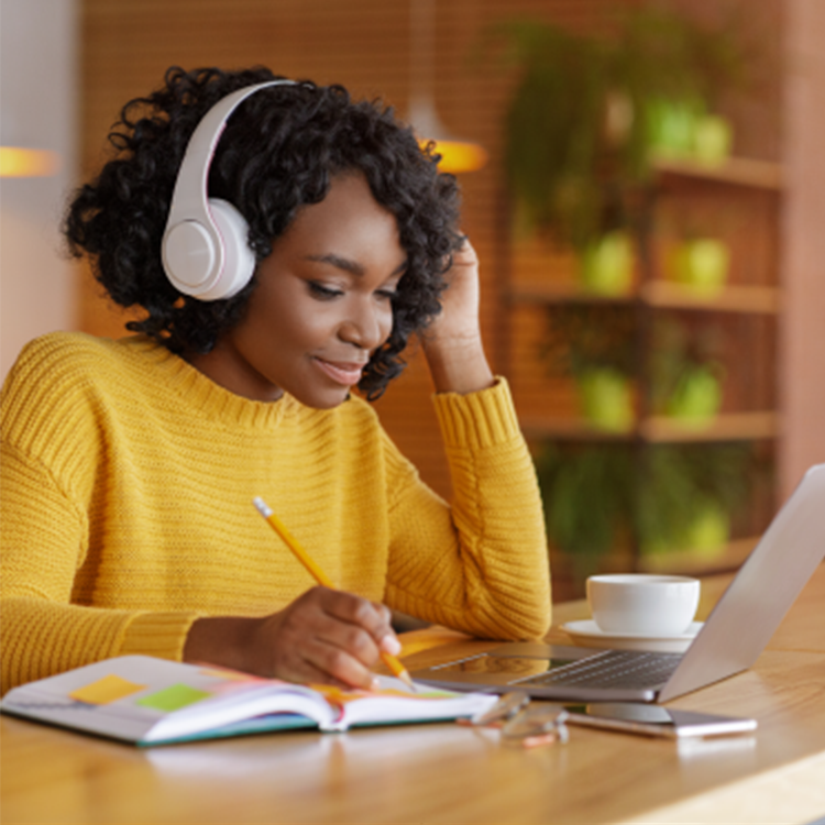 Imagem mostra uma mulher negra com fones de ouvido, sentada à frente de um notebook e com um lápis à mão. Na mesa, vemos um caderno de anotações, post-its e uma xícara.