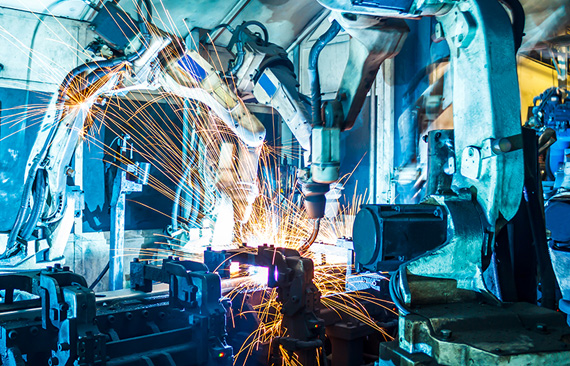 A imagem mostra uma fábrica de automóveis, na qual é possível ver mãos mecânicas realizando um procedimento de solda