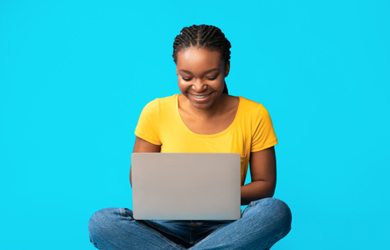 Mulher, sorrindo, utiliza notebook em seu colo. Ao fundo, cenário neutro todo azul.