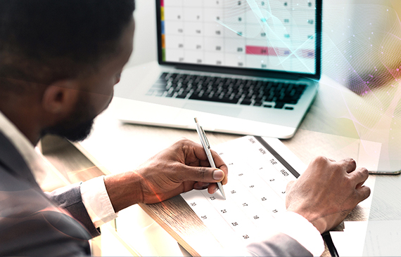 Imagem de um homem sentado observando um calendário na tela do seu laptop e fazendo anotações 