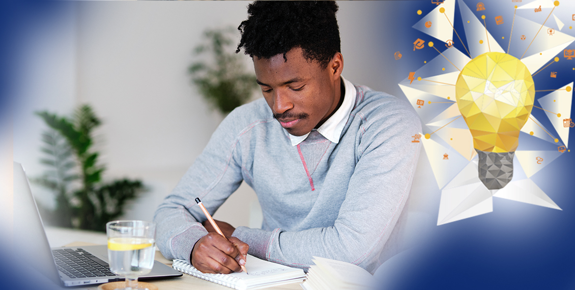 Homem escrevendo em um caderno, como se estivesse fazendo anotações do que está acompanhando no laptop que está aberto à sua frente. Ele está sentado em uma mesa