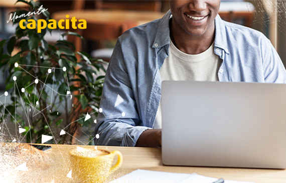 Imagem mostra um homem negro, de camisa azul, em frente a um computador portátil. Ele sorri e parece estudar, enquanto toma um café.