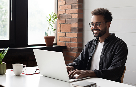 A imagem mostra um homem negro em frente a um computador. Ele veste uma camisa listrada e sorri.