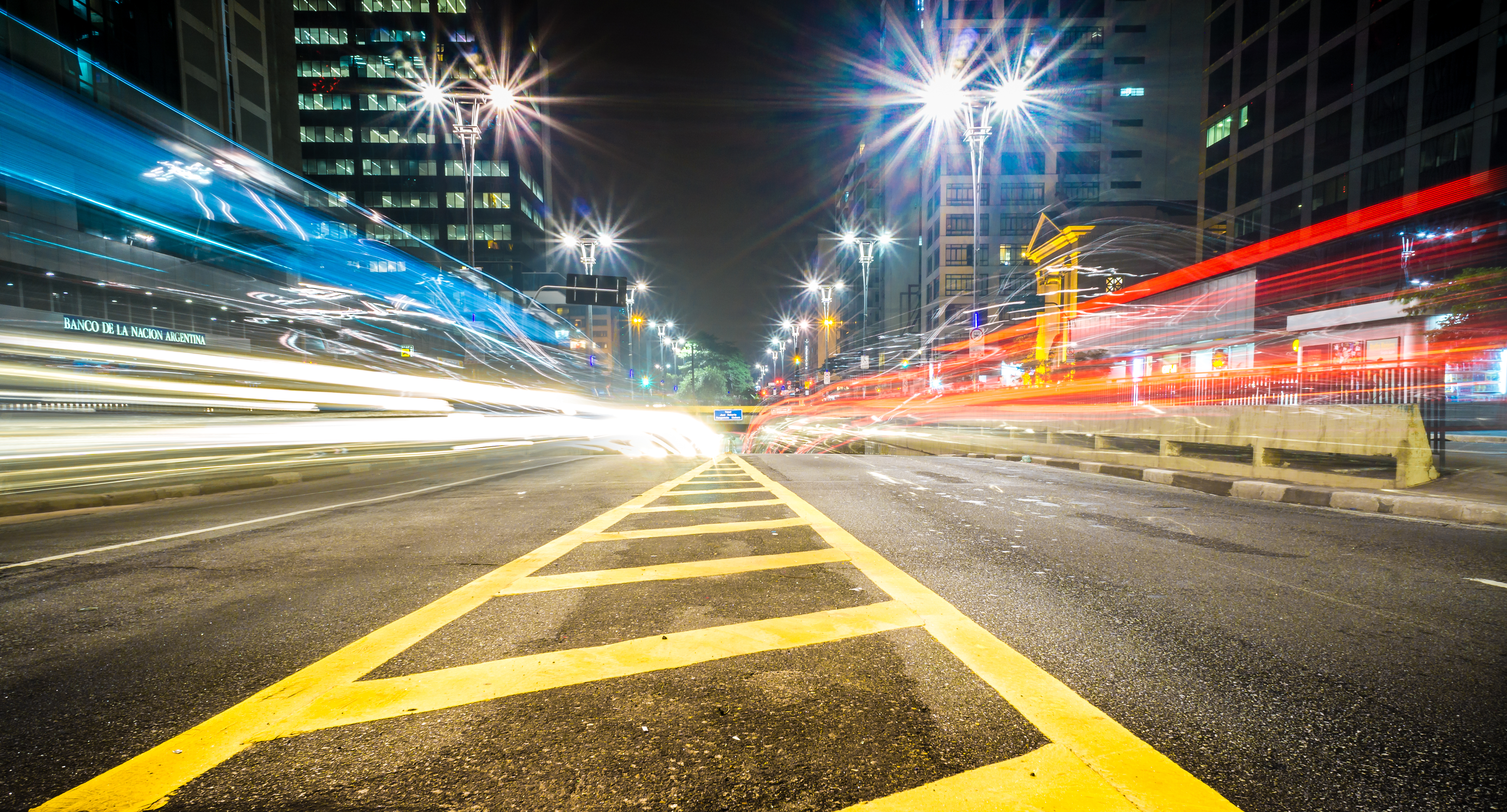 Imagem noturna de cidade mostra uma rodovia com postes de luz e carros em alta velocidade
