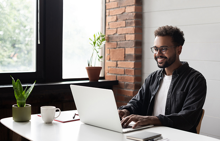 A imagem mostra um homem negro em frente ao seu computador. Ele veste uma camisa xadrez e sorri.