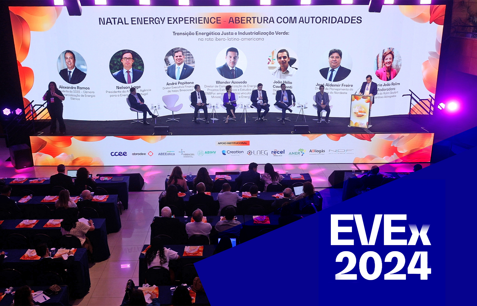 Foto do evento com pessoas sentadas e atrás um grande painel.