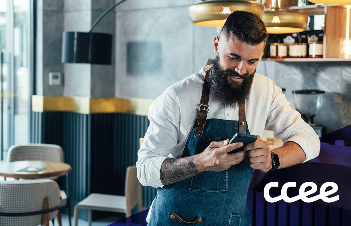 A imagem mostra um homem branco, de barba longa e preta, em um ambiente de comércio. Ele veste um avental e usa o seu celular enquanto sorri.