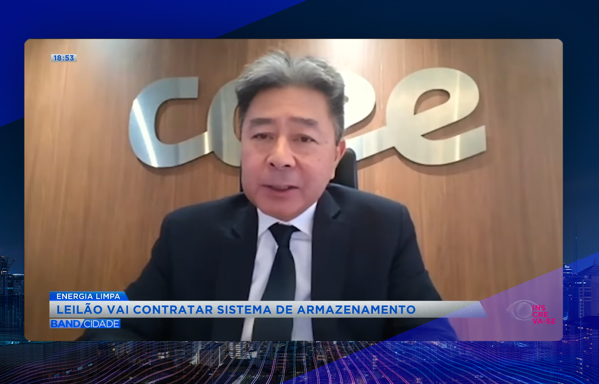A imagem mostra Ricardo Simabuku em frente a um painel de madeira, do qual se destaca o logo da CCEE. Ele é um homem asiático, de cabelos pretos, e veste um terno preto, camisa branca e gravata azul marinho.