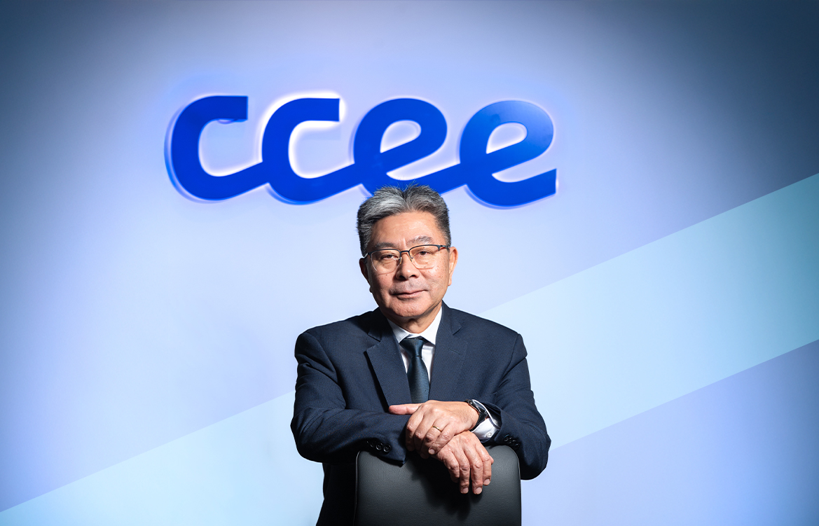 A imagem mostra o diretor-presidente interino da CCEE, Ricardo Simabuku, sentado em uma mesa de madeira. Ele é um homem asiático, de cabelos pretos e grisalhos. Veste um terno azul, com camisa branca e uma gravata preta. Atrás dele, está um letreiro com o logo da CCEE.