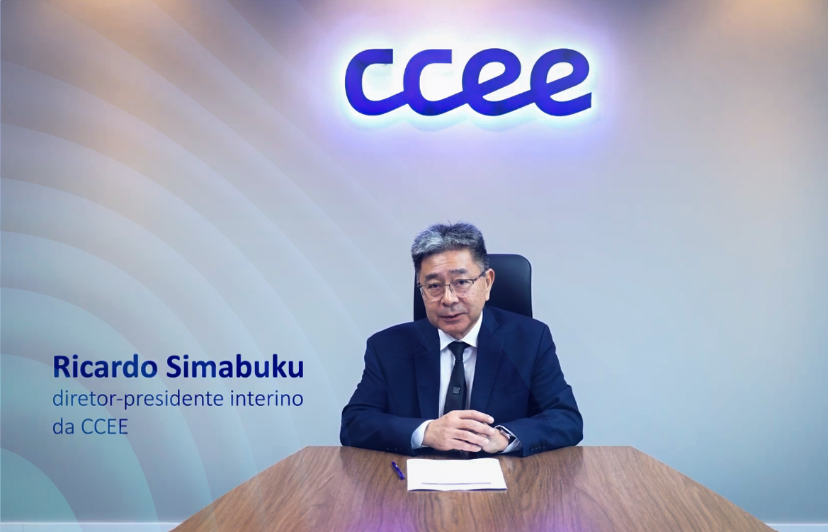 A imagem mostra o diretor-presidente interino da CCEE, Ricardo Simabuku, sentado em uma mesa de madeira. Ele é um homem asiático, de cabelos pretos e grisalhos. Veste um terno azul, com camisa branca e uma gravata preta. Atrás dele, está um letreiro com o logo da CCEE.