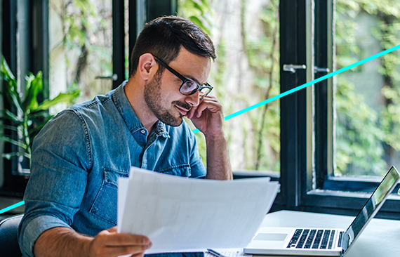 A imagem mostra um homem branco, de camisa azul, sentado em frente ao seu computador, em um cenário de escritório